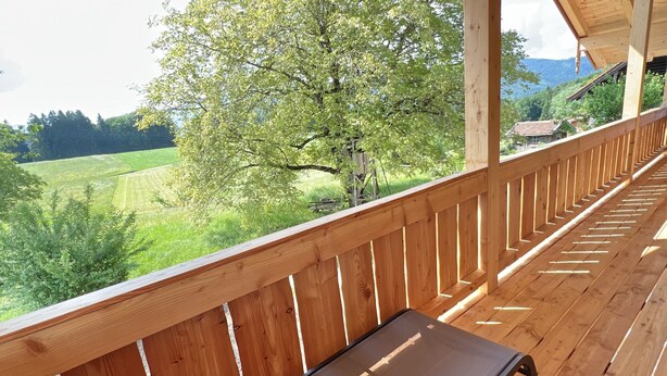 Una terraza de madera con un banco y una mesa, con vistas a un paisaje verde con árboles y una casa a lo lejos.