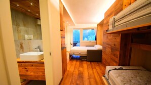 A narrow room with wooden walls and floors, a bed, a small bench, a window, and a sink.
