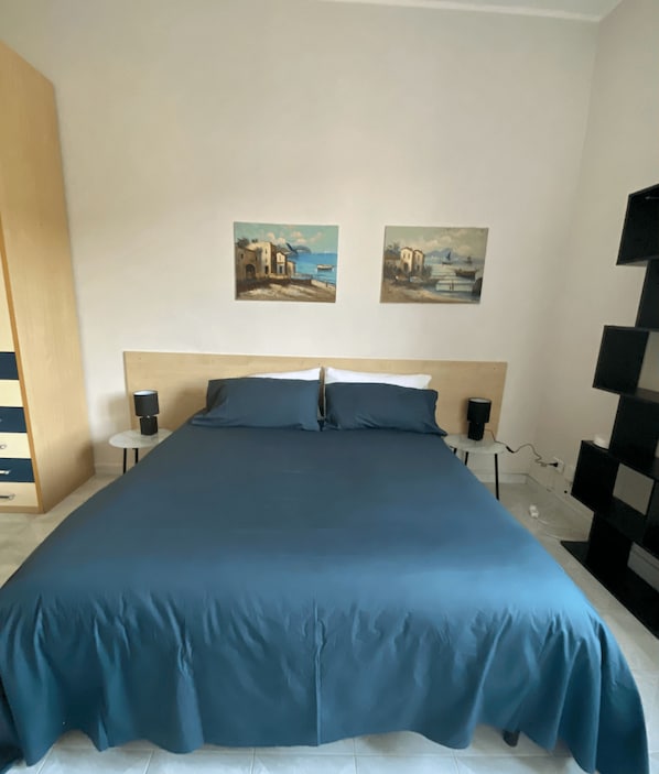 A bedroom with a large bed, two wall-mounted paintings, bedside lamps, and a black shelving unit.
