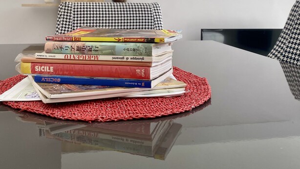 A neatly arranged stack of books on a glossy black table with a red woven placemat, a white wall with a wooden shelf holding decorative items, and a black chair in the background. - 5 minutes from the Valley of the Temples, apartment with free parking (Agrigento)