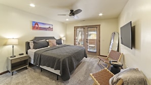 A bedroom with a bed, bedside table, lamp, chair, TV, ceiling fan, and a door leading to a balcony. - Red Rock Oasis I Large Patio I Pool & Hot Tub I Views (Moab)