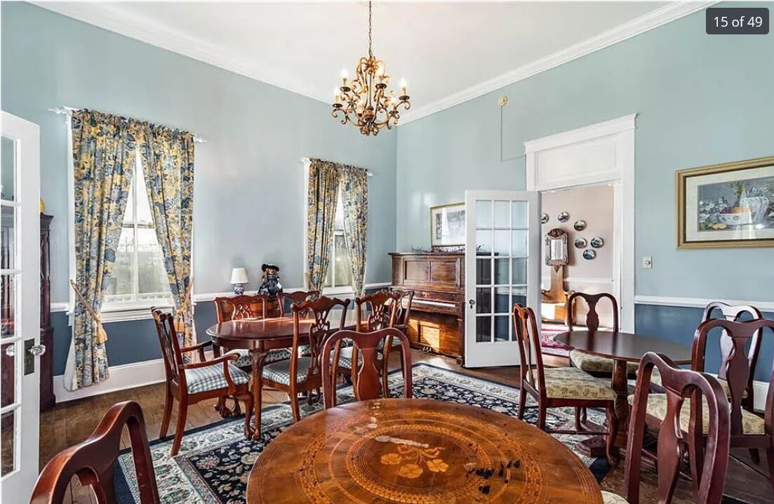 Een eetkamer met een ronde houten tafel, meerdere stoelen, een kroonluchter en bloemen gordijnen.