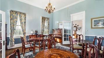 Een eetkamer met een ronde houten tafel, meerdere stoelen, een kroonluchter en bloemen gordijnen.