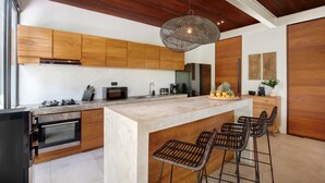 A modern kitchen with wooden cabinets, a large island with bar stools, a hanging light fixture, and a built-in oven.