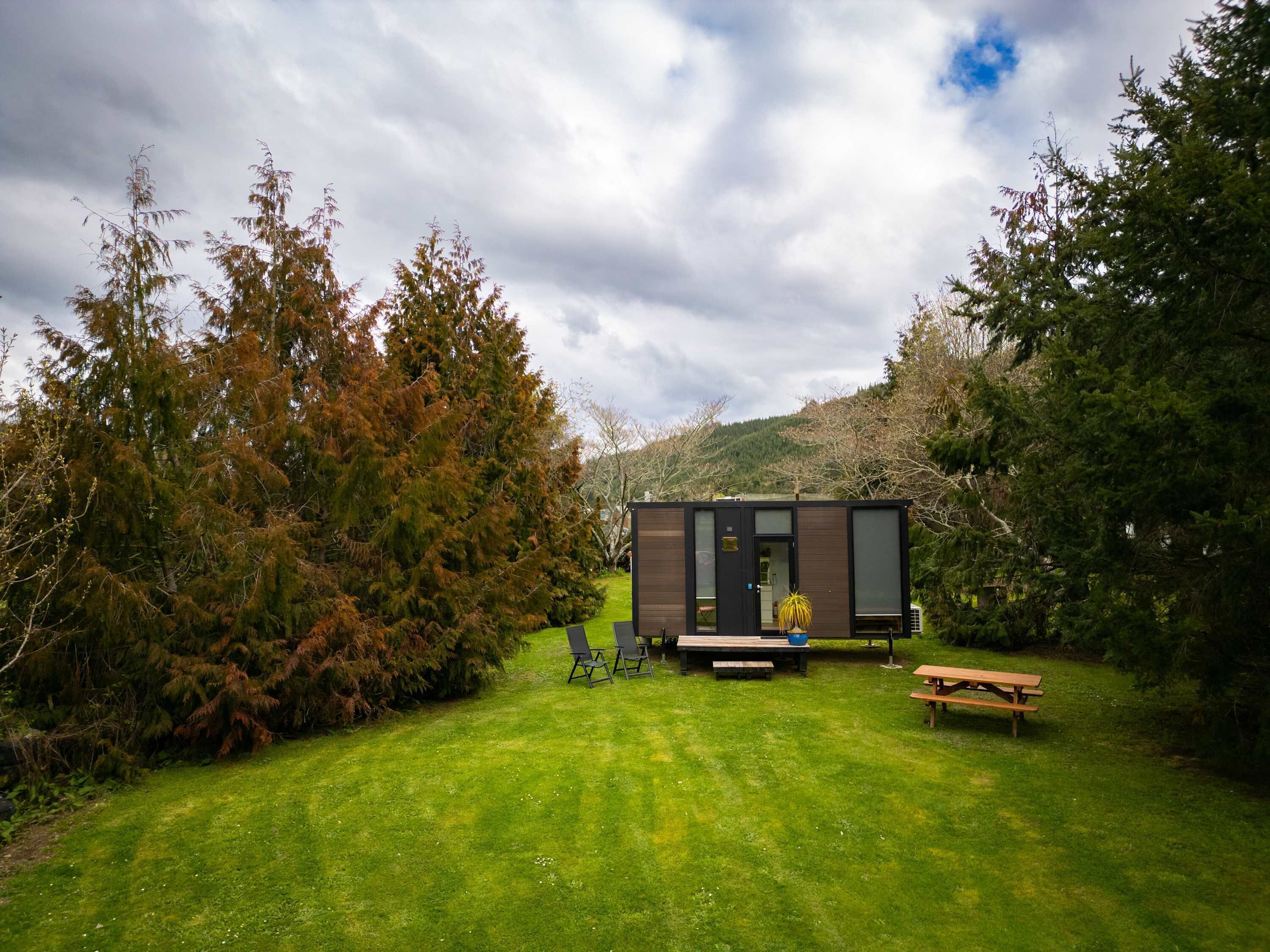 A small cabin with a deck, surrounded by trees and a grassy lawn.