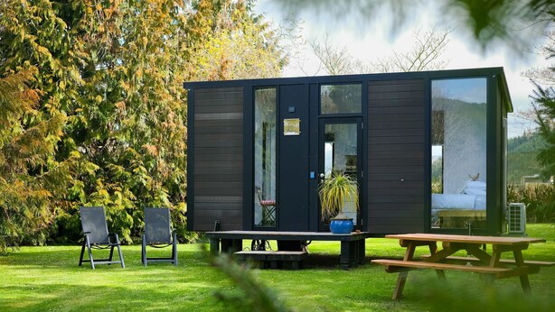 A modern cabin with a large glass door, a bench, two chairs, and a picnic table in a grassy area with trees. - Ryans Ranch by Tiny Away (Belgrove)