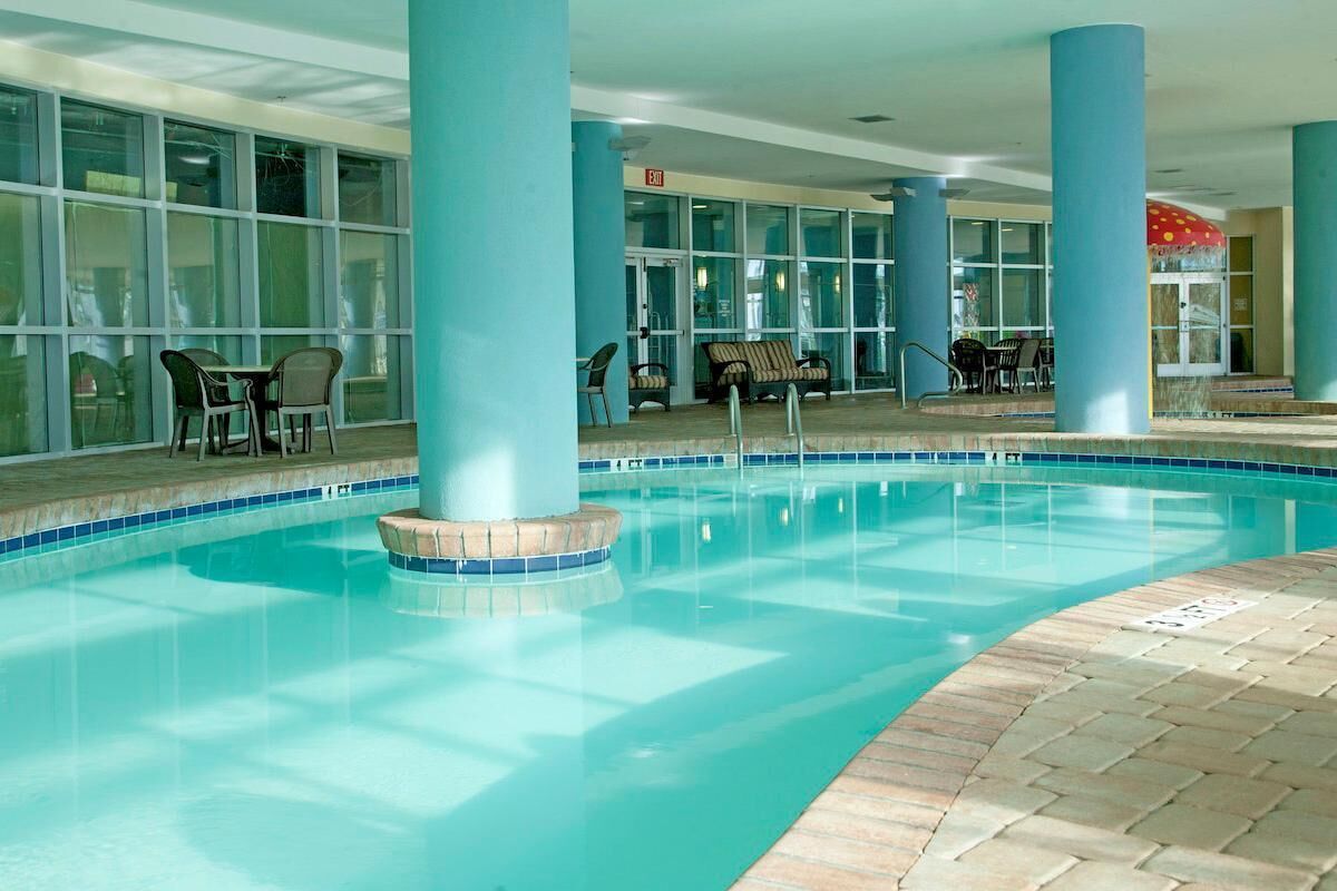 An indoor swimming pool with clear blue water, surrounded by blue pillars and a tiled deck. - Shoreline Serenity: King Bed, Private Balcony (Myrtle Beach)