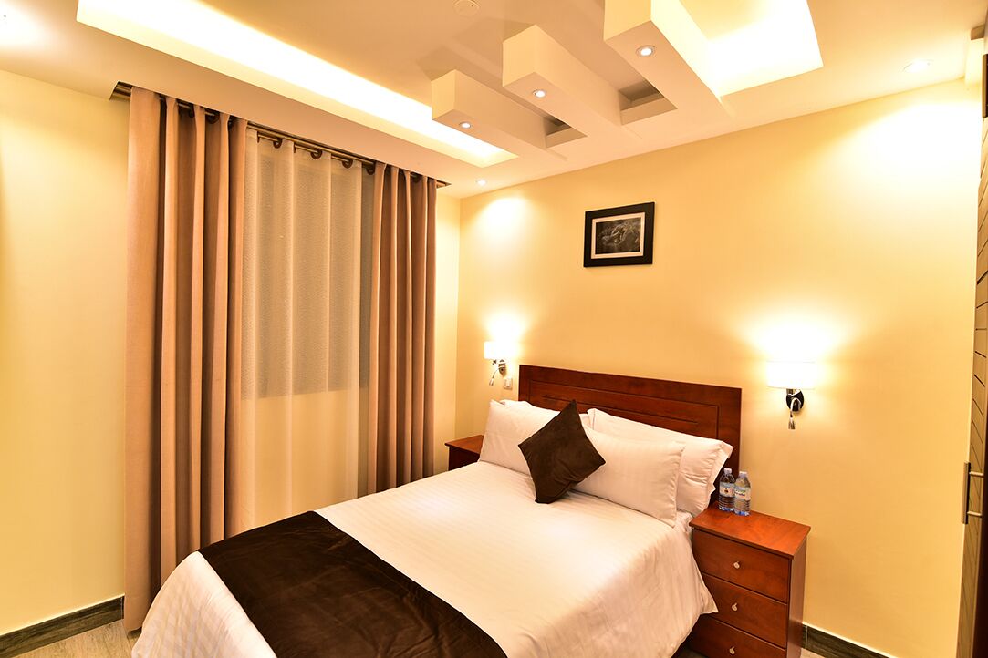 Una habitación de hotel con cama, cortinas, mesita de noche e iluminación en la pared.