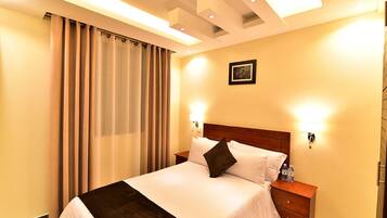 Una habitación de hotel con cama, cortinas, mesita de noche e iluminación en la pared.