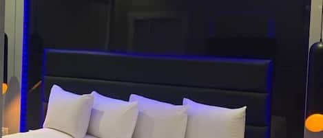 Une chambre moderne avec un grand lit, une tête de lit sombre et un éclairage d’ambiance bleu.