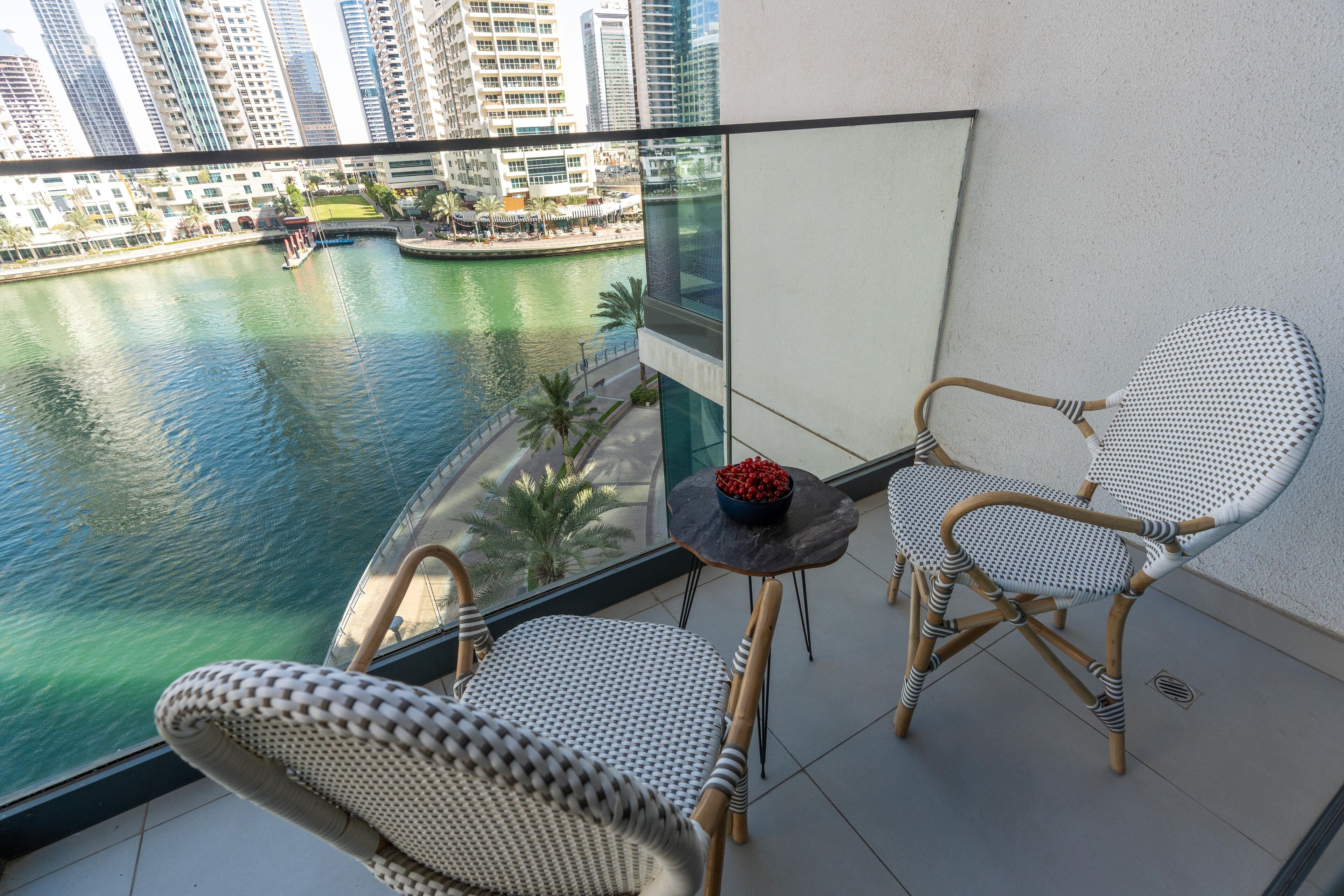 A balcony with wicker chairs and a small table, overlooking a body of water with buildings in the background.