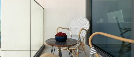 A balcony with wicker chairs, a small round table with a bowl of fruit, and a large window.