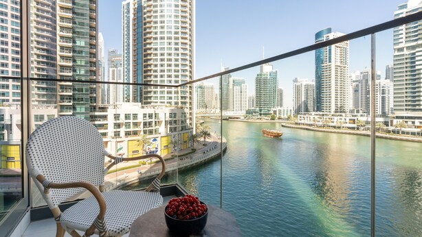 A balcony with a wicker chair and a small table with a bowl of fruit, overlooking a river with boats and high-rise buildings in the background. - LIV Residence (Dubai)
