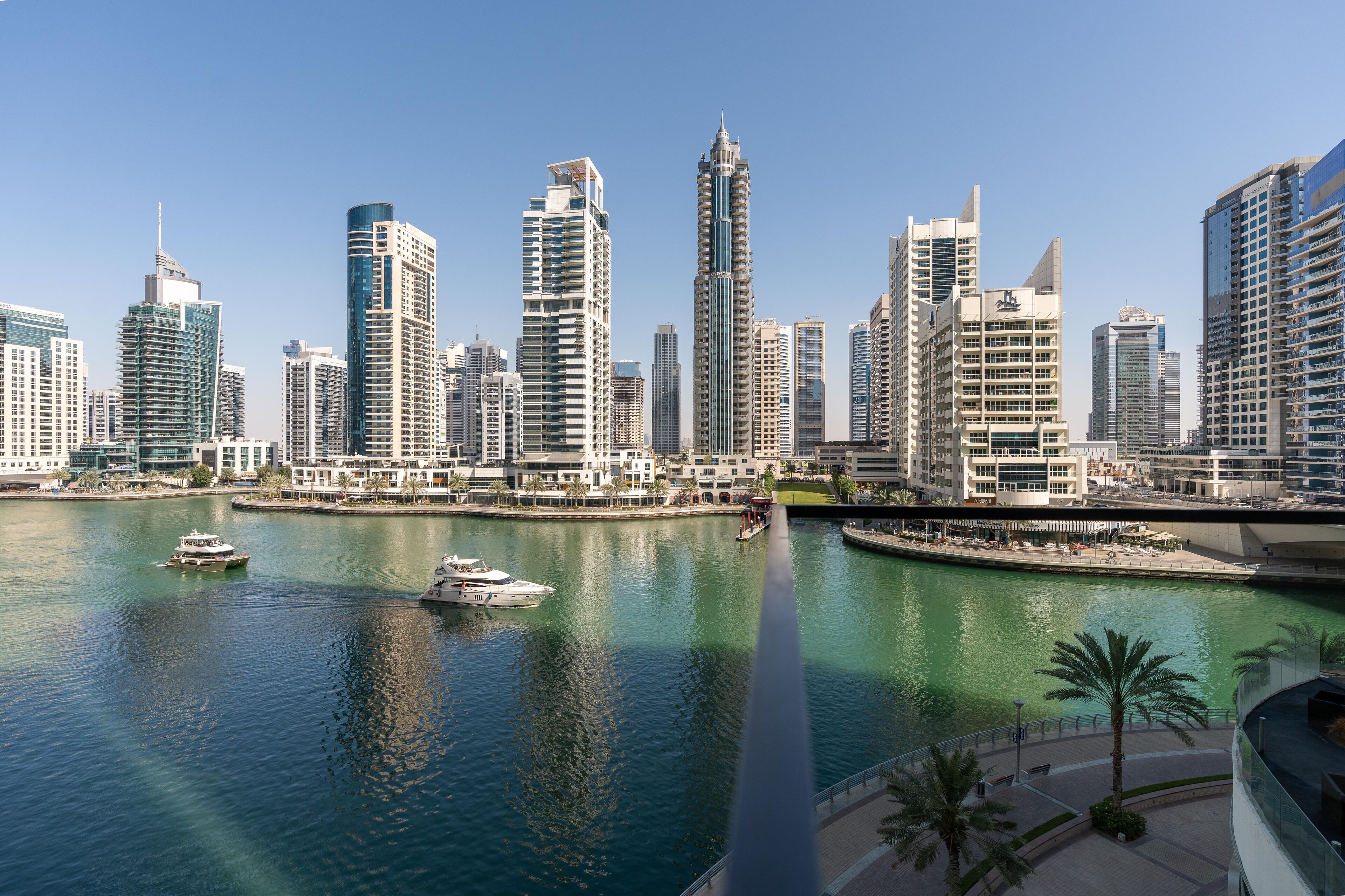 A cityscape with modern high-rise buildings along a waterfront, a boat on the water, and a pedestrian walkway with palm trees.