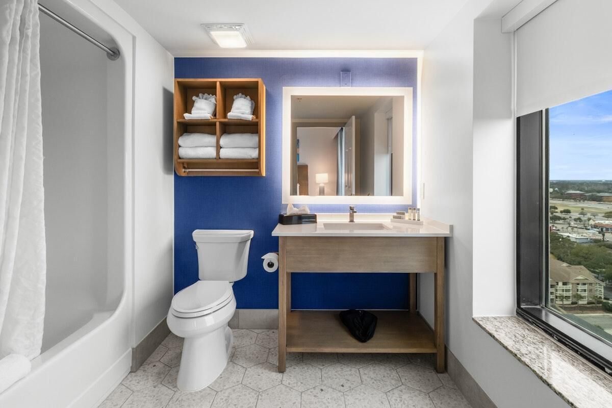Un bagno moderno con il water bianco, un mobile lavabo in legno con specchio e vista sulla città attraverso un'ampia finestra.