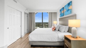 A hotel room with a large bed, a nightstand, a lamp, and a view of the city through a large window. - Newly Renovated Ocean View 3 Bdrm Condo 6th Flr (Myrtle Beach)