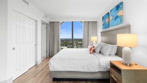 A hotel room with a large bed, a nightstand, a lamp, and a view of the city through a large window.