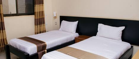 A hotel room with two single beds, a window with curtains, a bedside table, and a nightstand.