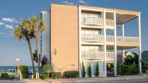 Un immeuble à plusieurs étages doté de balcons, offrant une vue sur la plage et comprenant un espace de stationnement agrémenté de palmiers.