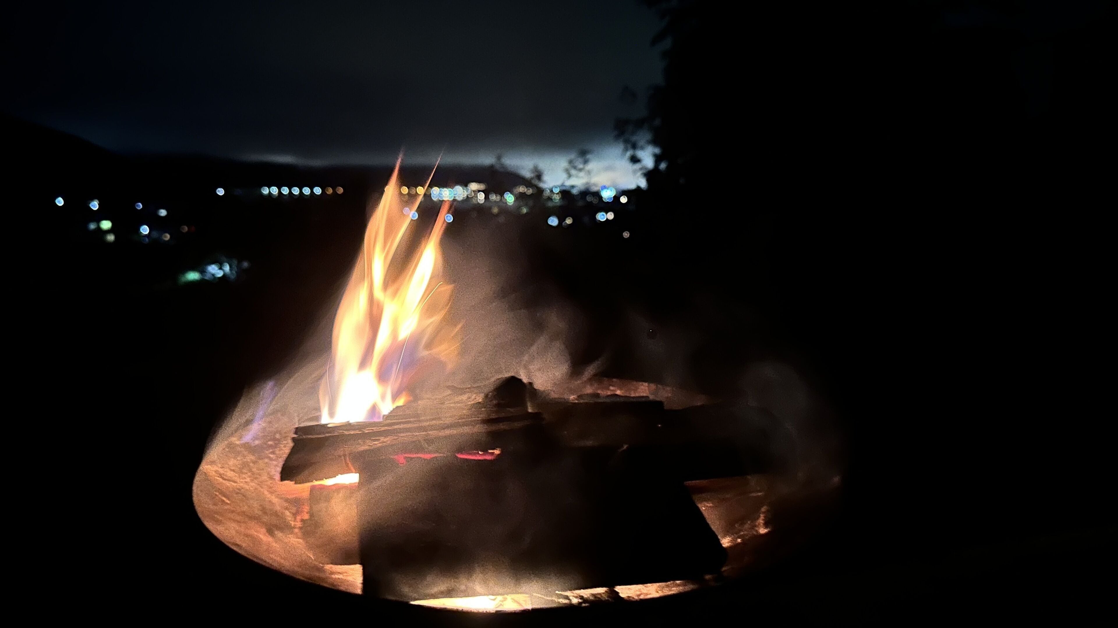 Hoguera con troncos y un recipiente metálico, rodeada de un área boscosa oscura.