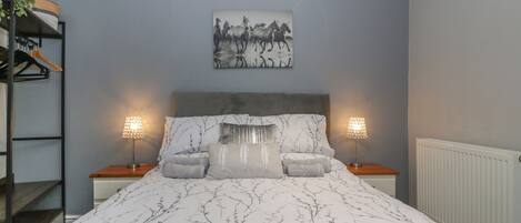 A bedroom with a bed, two bedside lamps, a headboard, a wall-mounted painting, and a radiator.