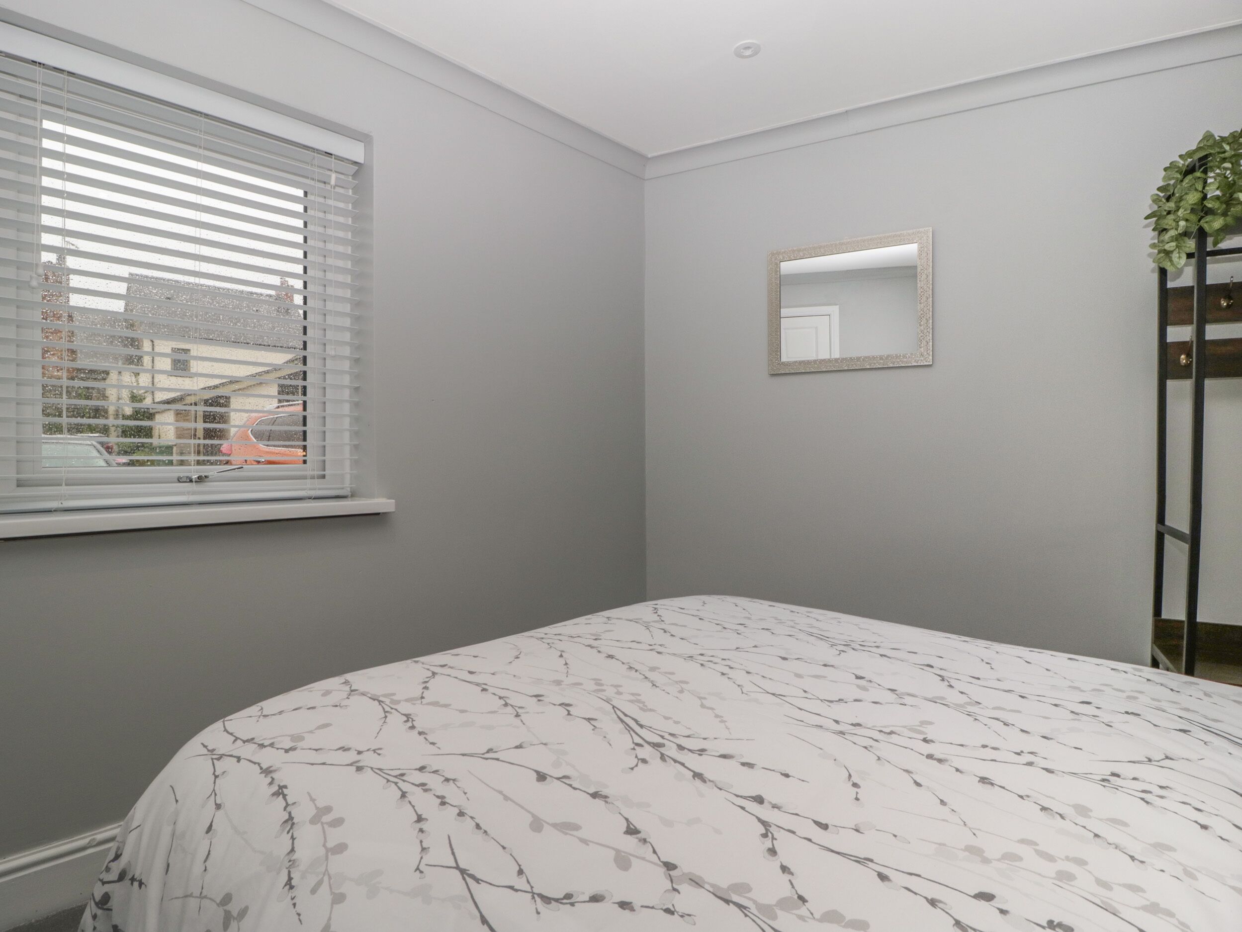 Ein Schlafzimmer mit einem Bett, einem Fenster mit Jalousien, einem Spiegel und einer Pflanze.