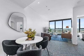 A modern hotel room with a dining area, a sofa, a TV, and a large window offering a city view.