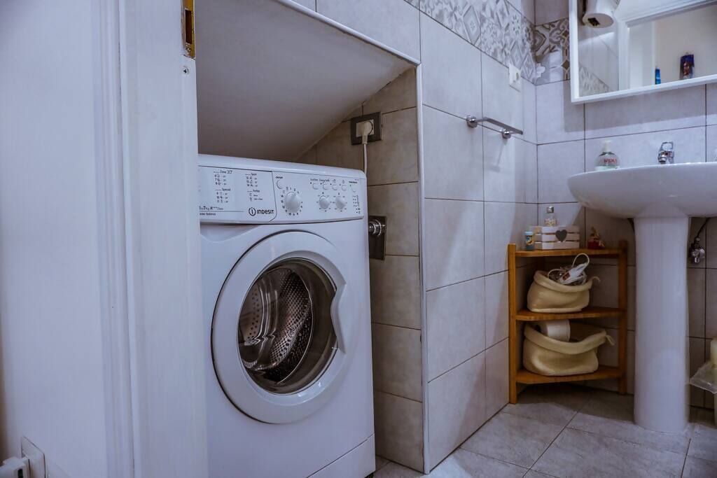 A compact laundry room with a front-loading washing machine, a sink, and a shelf with bags.