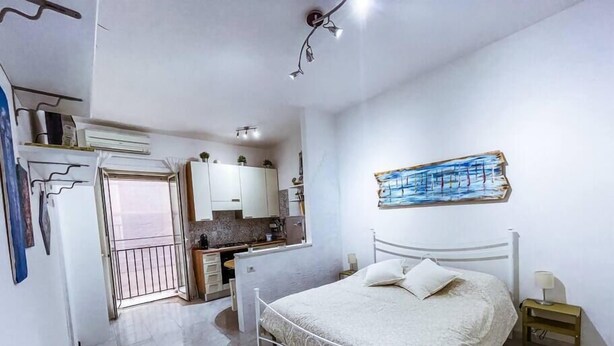 A bedroom with a bed, a nightstand, a wall-mounted painting, and a kitchenette in the background.