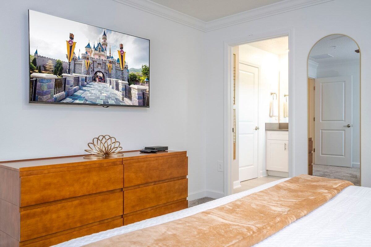 Une chambre avec un grand lit, une commode en bois, un téléviseur à écran plat fixé au mur et une porte donnant sur une autre pièce.