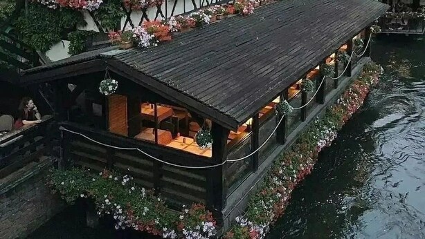 Un edificio tradizionale con struttura a telaio in legno e numerosi balconi decorati con fiori, un pontile in legno che si estende su una distesa d'acqua, con un fiume che scorre accanto.