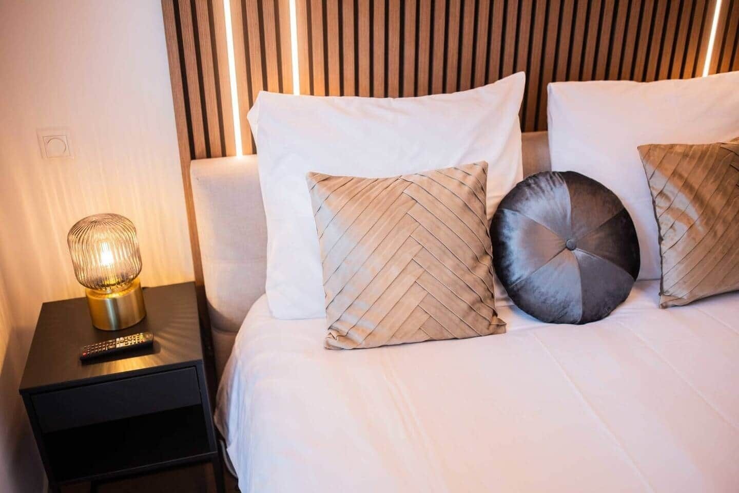 Una cama bien hecha con ropa blanca y tres almohadones decorativos, una lámpara de noche y un control remoto sobre una mesita de noche negra.