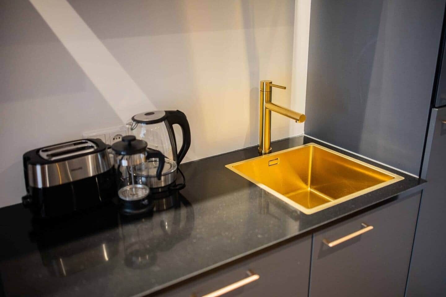 A kitchen with a black countertop, a stainless steel toaster, a glass coffee maker, and a gold-colored sink.