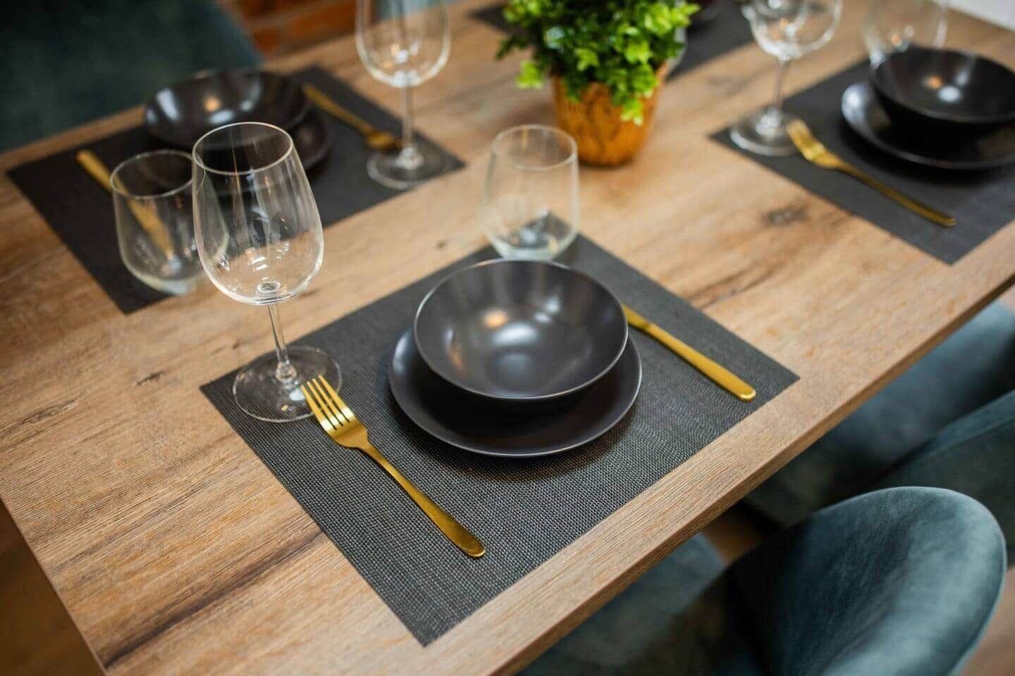 Une table à manger dressée avec des assiettes noires, des couverts dorés et des verres à vin, le tout posé sur une table en bois surmontée d’un chemin de table foncé.