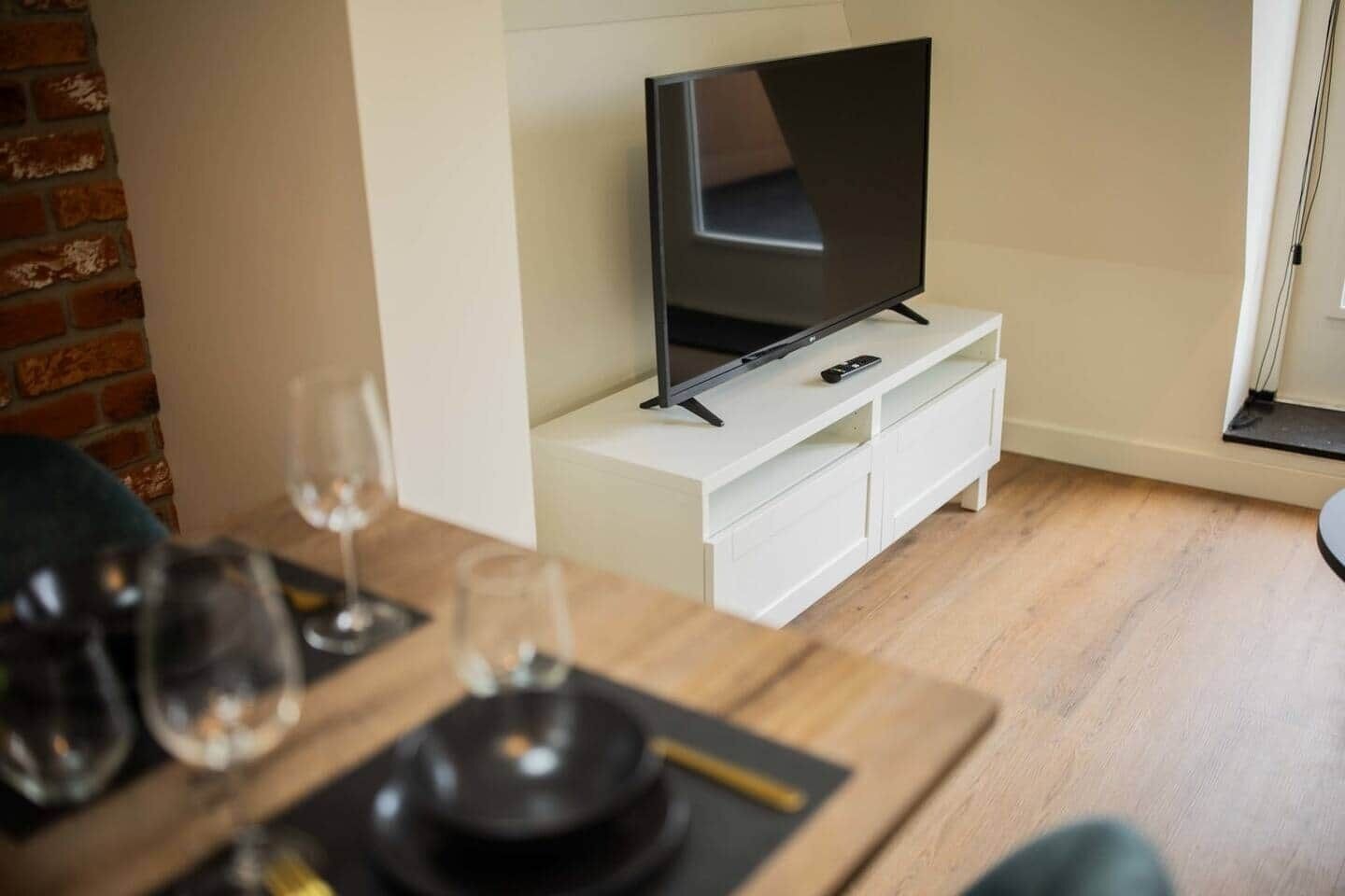 Una sala moderna con una televisión de pantalla plana sobre un mueble blanco, una mesa de comedor con vasos y cubiertos, y una pared de ladrillo.