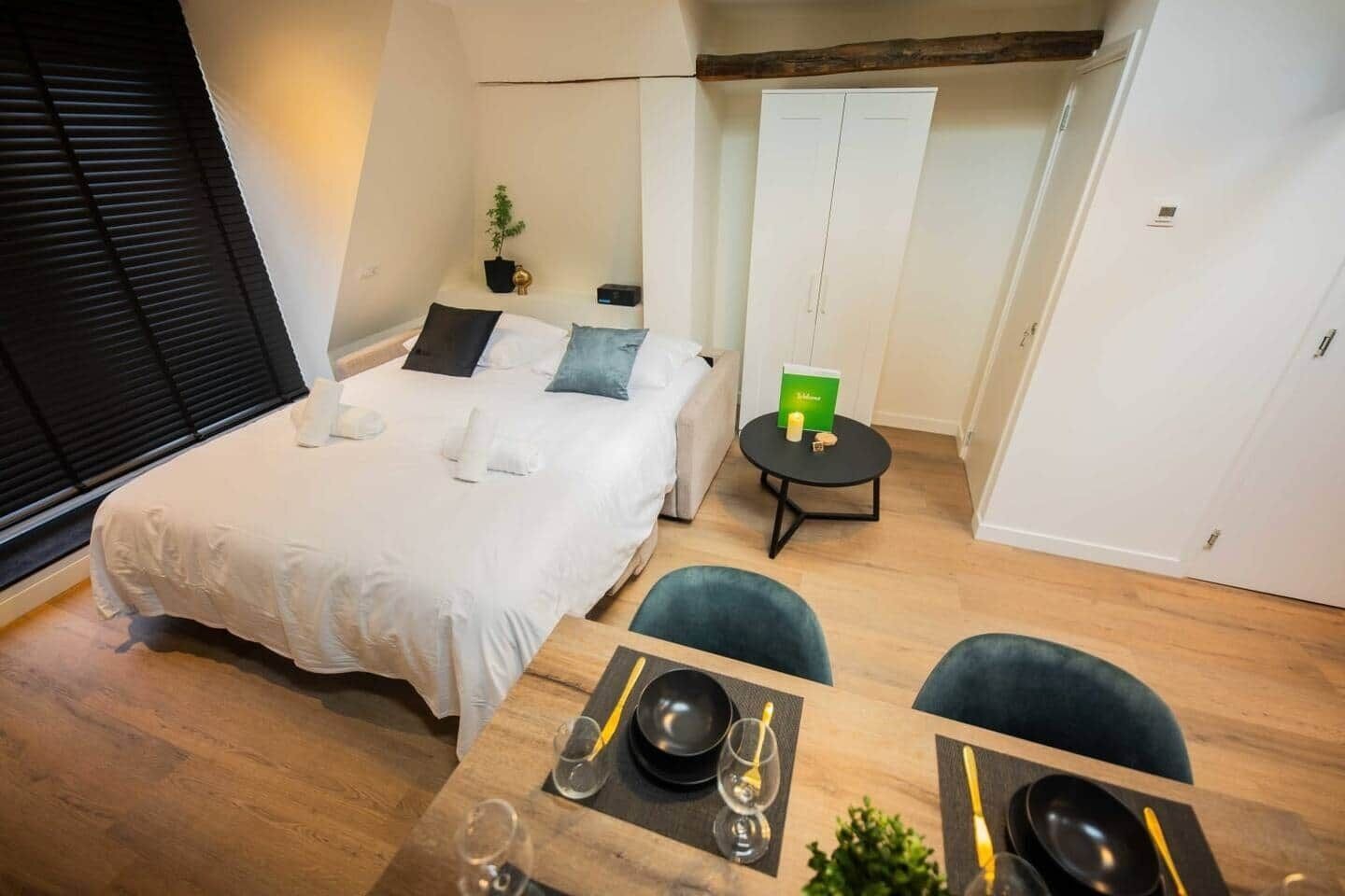 A hotel room with a bed, a small dining table with chairs, a round table with a book and candle, and a wardrobe.