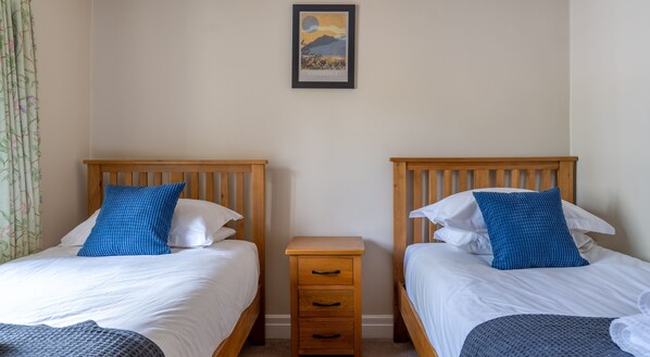 A room with two wooden beds, each with a blue pillow and white bedding. There is a small wooden nightstand with drawers between the beds. A framed picture hangs on the wall above the beds. - Bassicks - sleeps 8 guests in 4 bedrooms (Graythwaite)