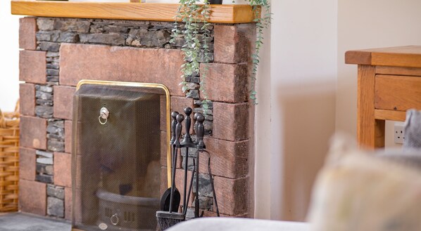 A stone fireplace with a metal grate, a wooden mantel, and a hanging plant. - Bibby Lot - sleeps 6 guests  in 3 bedrooms (Graythwaite)