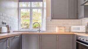 A kitchen with grey cabinets, a window with a view of greenery, a bread container, a toaster, a kettle, and a microwave.
