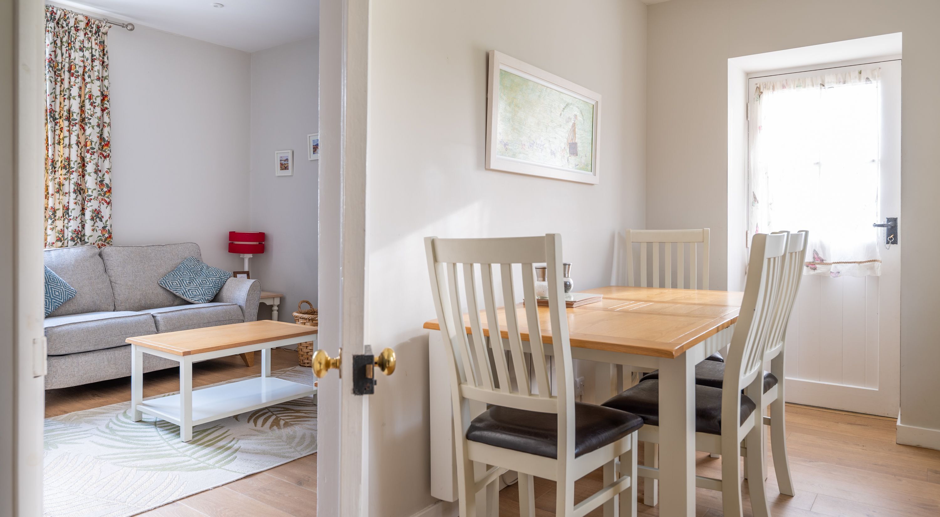 Un coin repas avec une table en bois et des chaises blanches, un canapé agrémenté de coussins à motifs, une lampe rouge et une porte avec un rideau blanc.