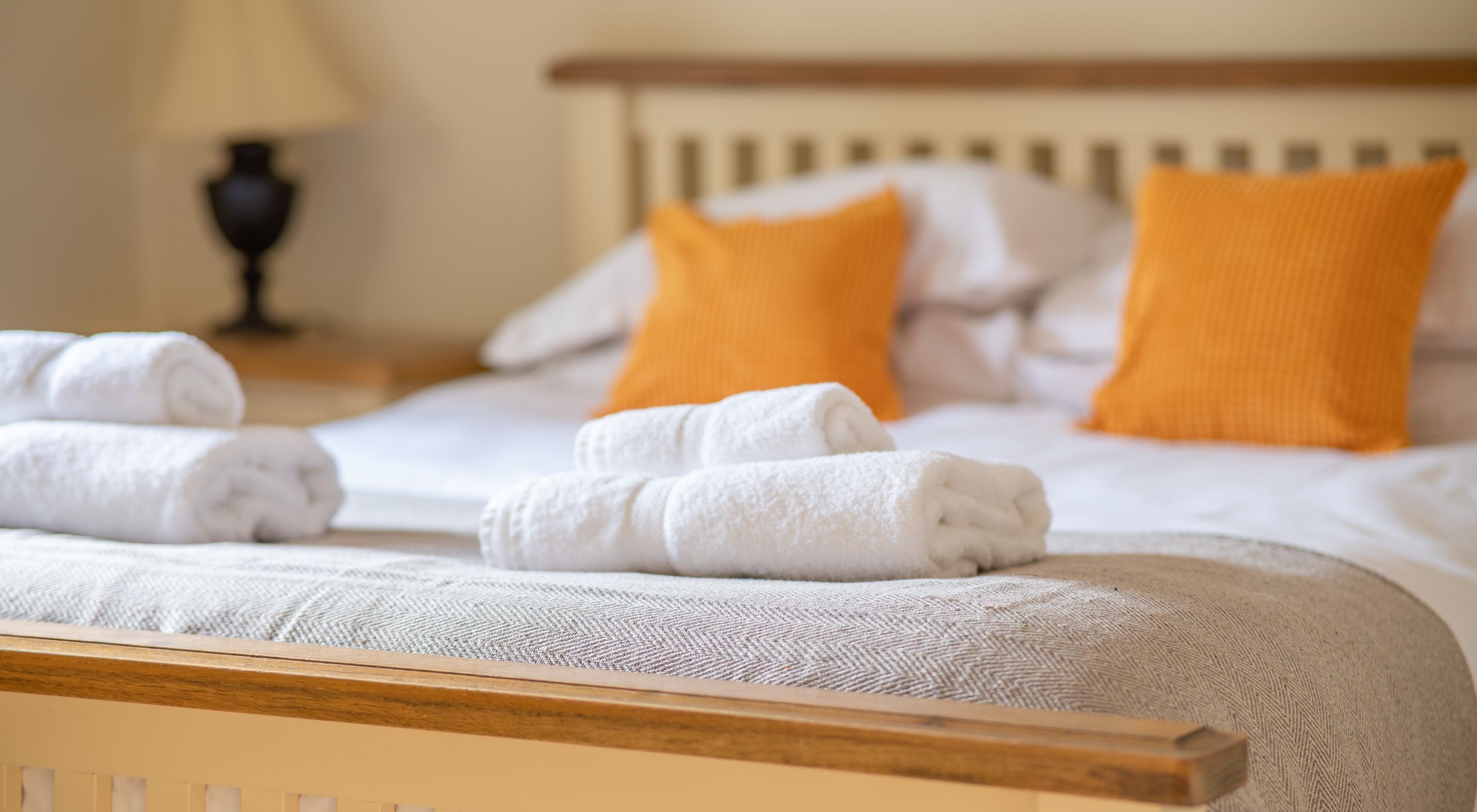 Un lit bien fait, avec du linge de lit blanc et deux coussins orange, une tête de lit en bois et une lampe en arrière-plan.