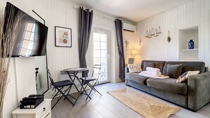 A living room with a grey sofa, a small table with chairs, a flat-screen TV, and a large window with curtains. - (058) Le petit National (Antibes)