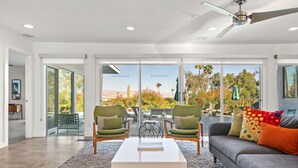Un salon moderne avec une table basse blanche, deux fauteuils verts, un canapé gris agrémenté de coussins colorés et une vue sur un jardin, visible à travers de grandes portes vitrées.