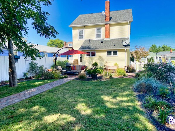 Una casa de dos pisos con un patio frontal, un patio trasero con mobiliario exterior y una cerca blanca de piquetes.