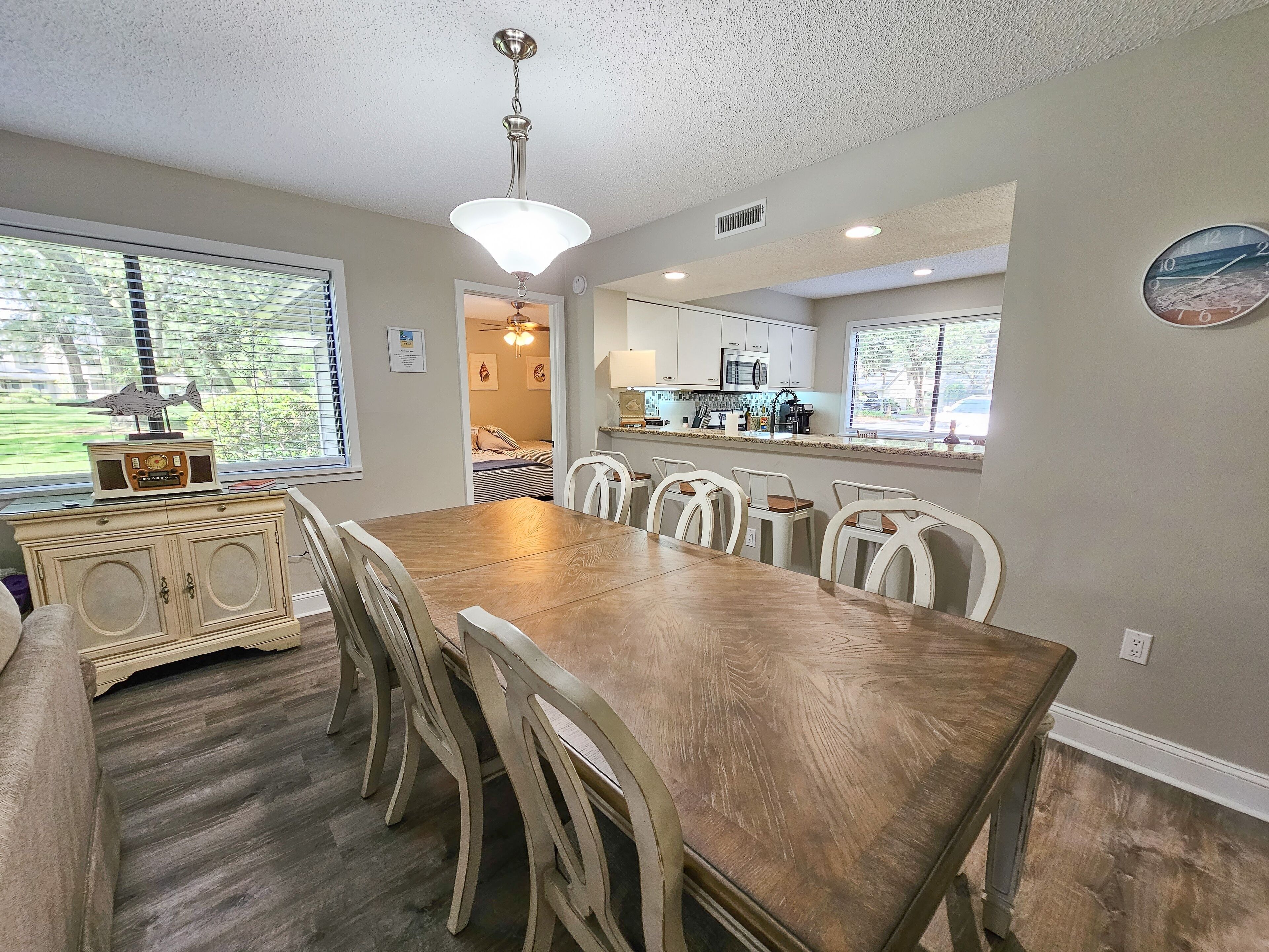 A dining area with a wooden table and chairs, a ceiling fan, and a view of the outdoors through large windows.
