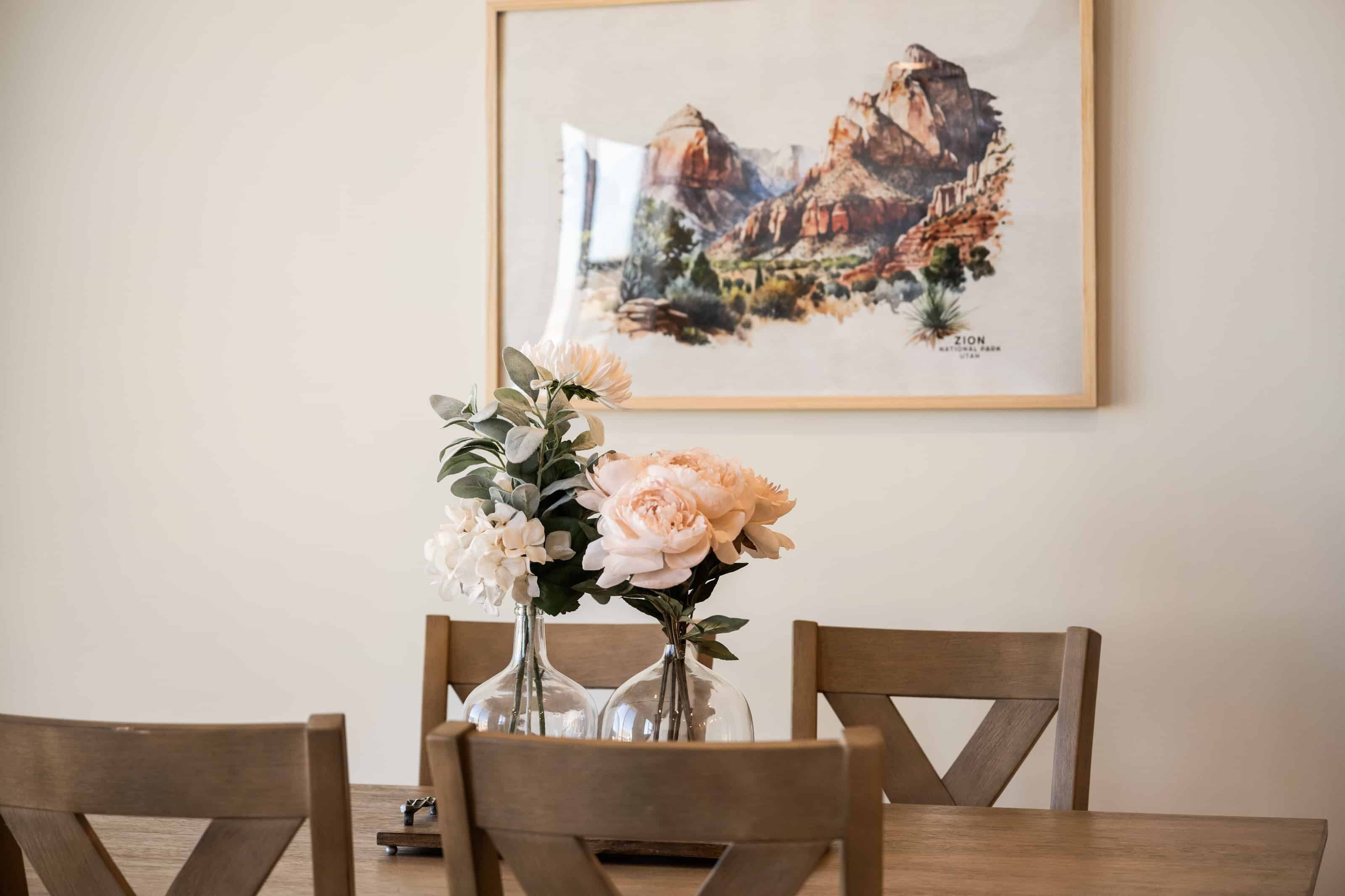 A dining table with two glass vases of flowers, wooden chairs, and a framed painting on the wall.