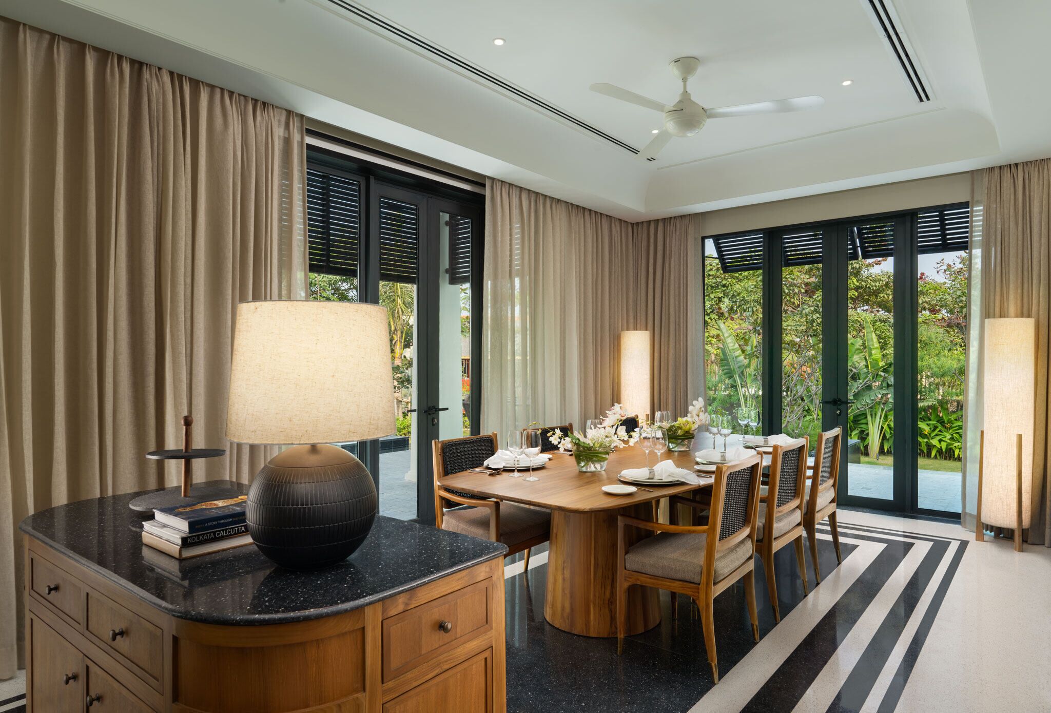 A dining area with a wooden table set for four, surrounded by chairs, with a large window offering a view of greenery.
