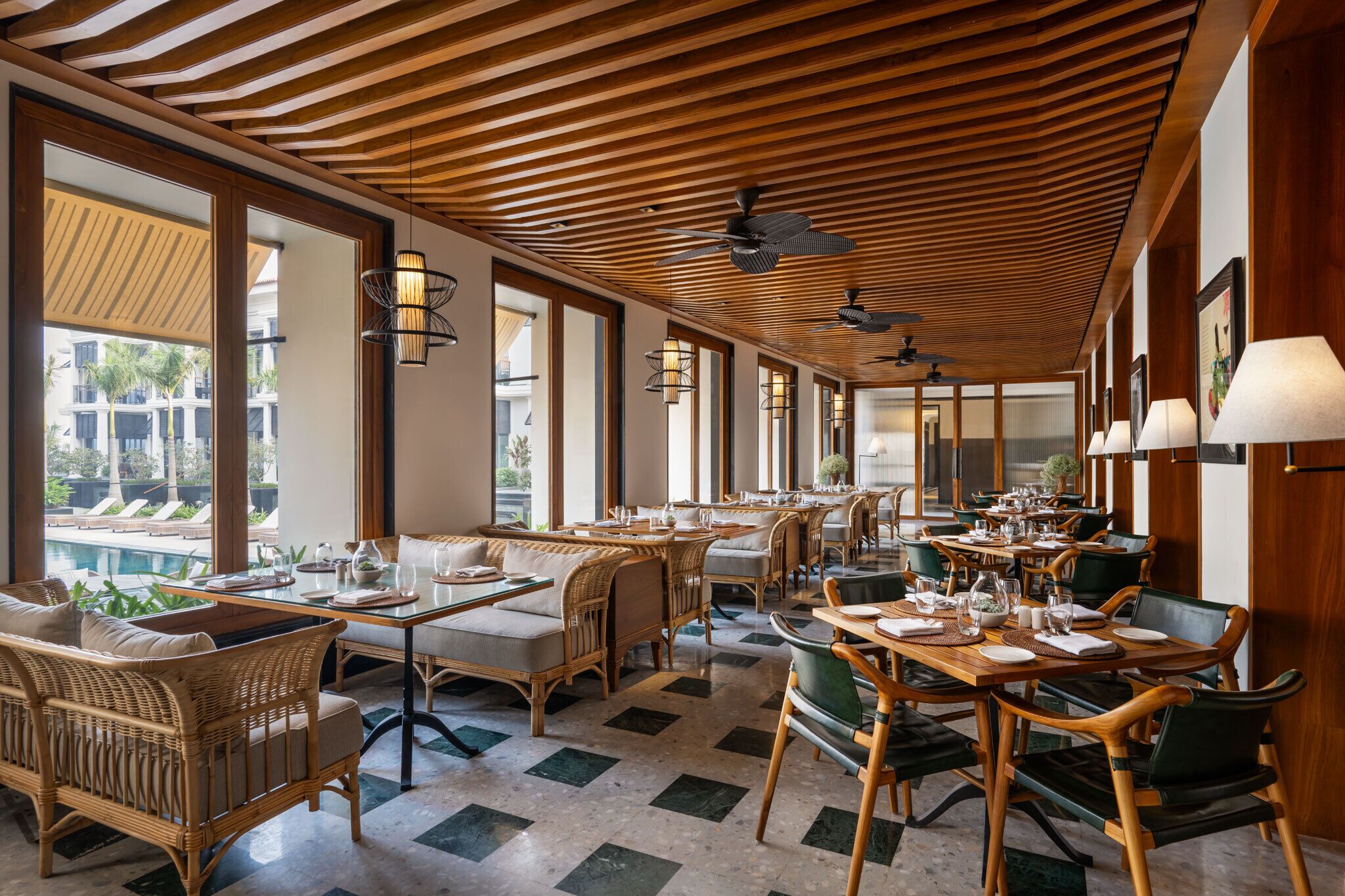 A spacious indoor dining area with wooden ceiling beams, large windows, and a pool view. Tables are set with chairs and dining ware.