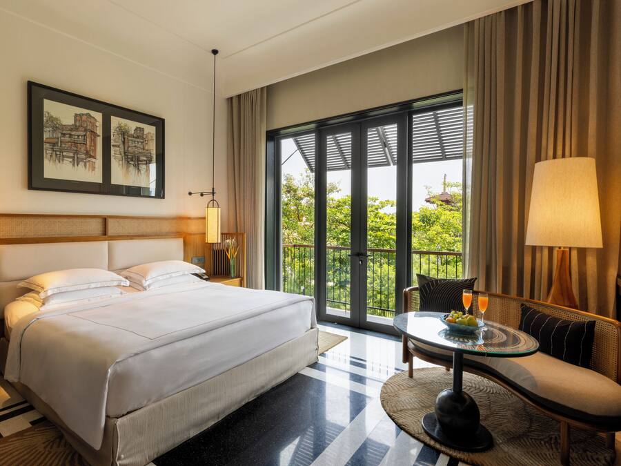 A hotel room with a large bed, a sofa, a small table with fruit and drinks, and a balcony with a view of greenery.