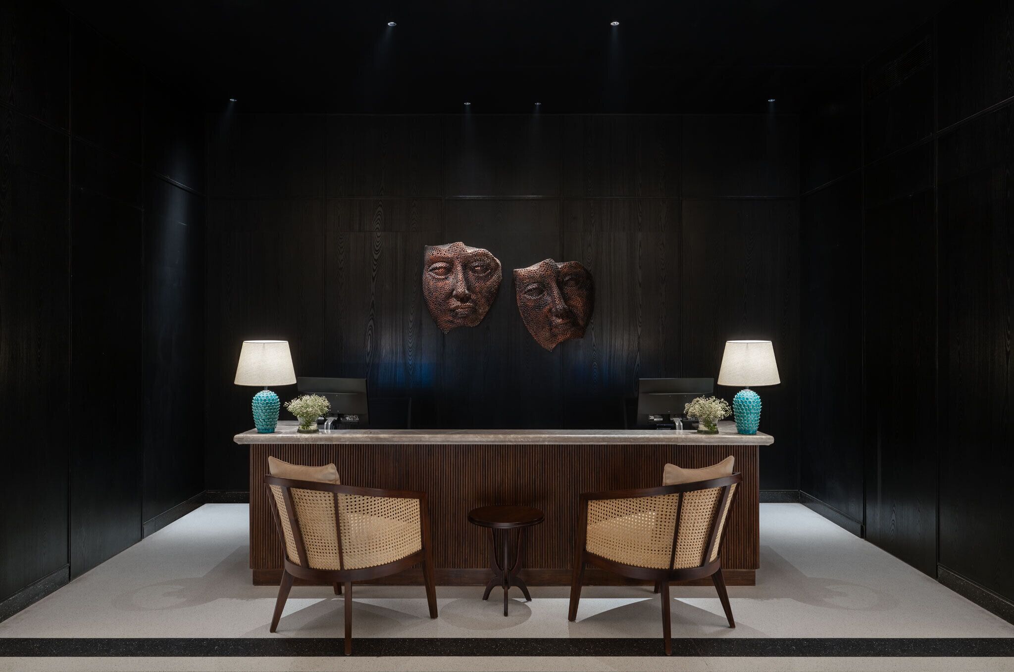 Zona de recepción con pared de madera oscura, dos máscaras colgadas en la pared, un mostrador de recepción, dos sillas de mimbre y dos lámparas.